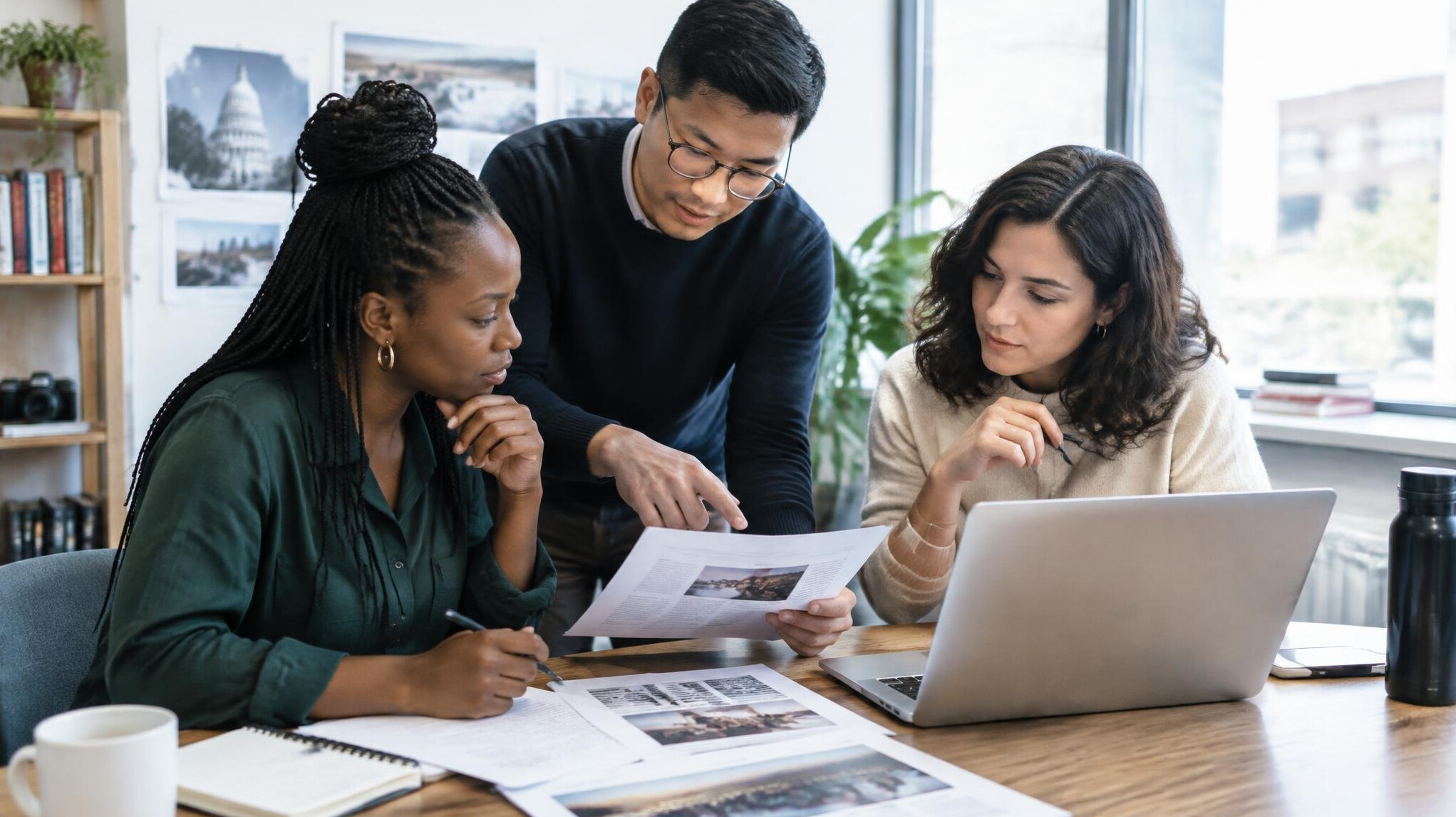 Small editorial team gathered around a table reviewing printed documents and a laptop